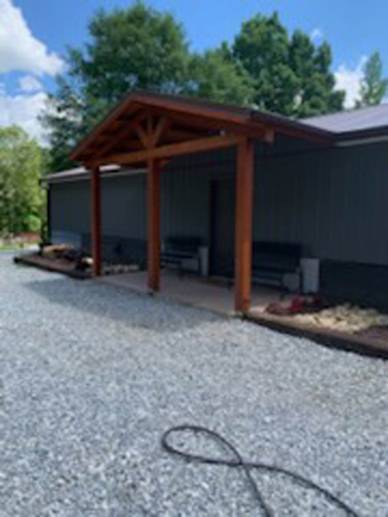 Gray building with brown wood porch, gravel driveway, and surrounding trees.