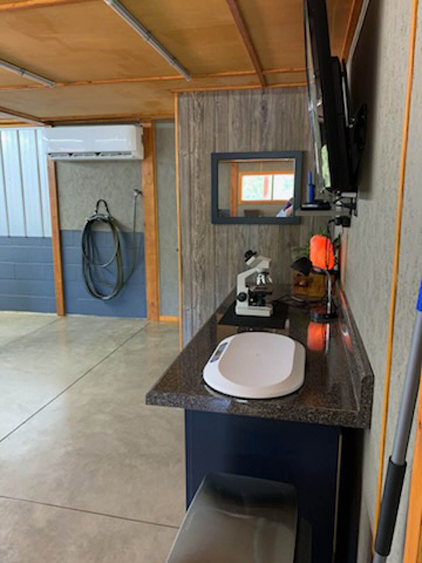 A counter with a microscope, scale, and TV. A hose and an AC unit are on the wall. The room has concrete flooring.