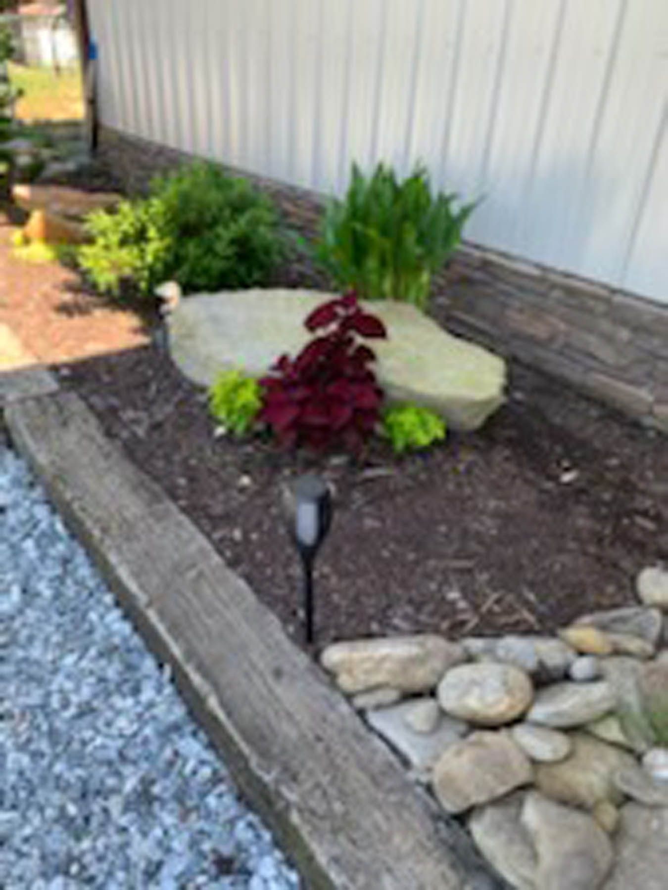 Flowerbed with rock border and stone accents, plants, and a solar light next to a white building.