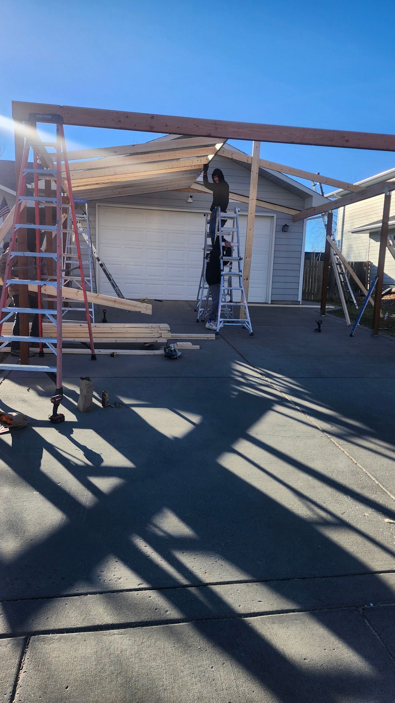 Construction workers building an outdoor structure; ladder, wooden beams, blue sky, garage in background.