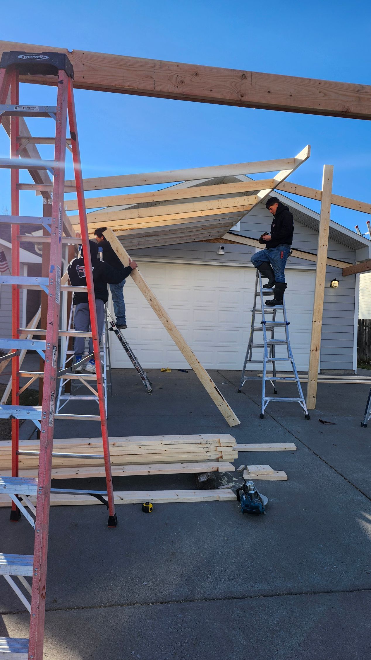 Construction workers building a wooden structure in front of a garage. One on a ladder, others holding boards. Clear sky.