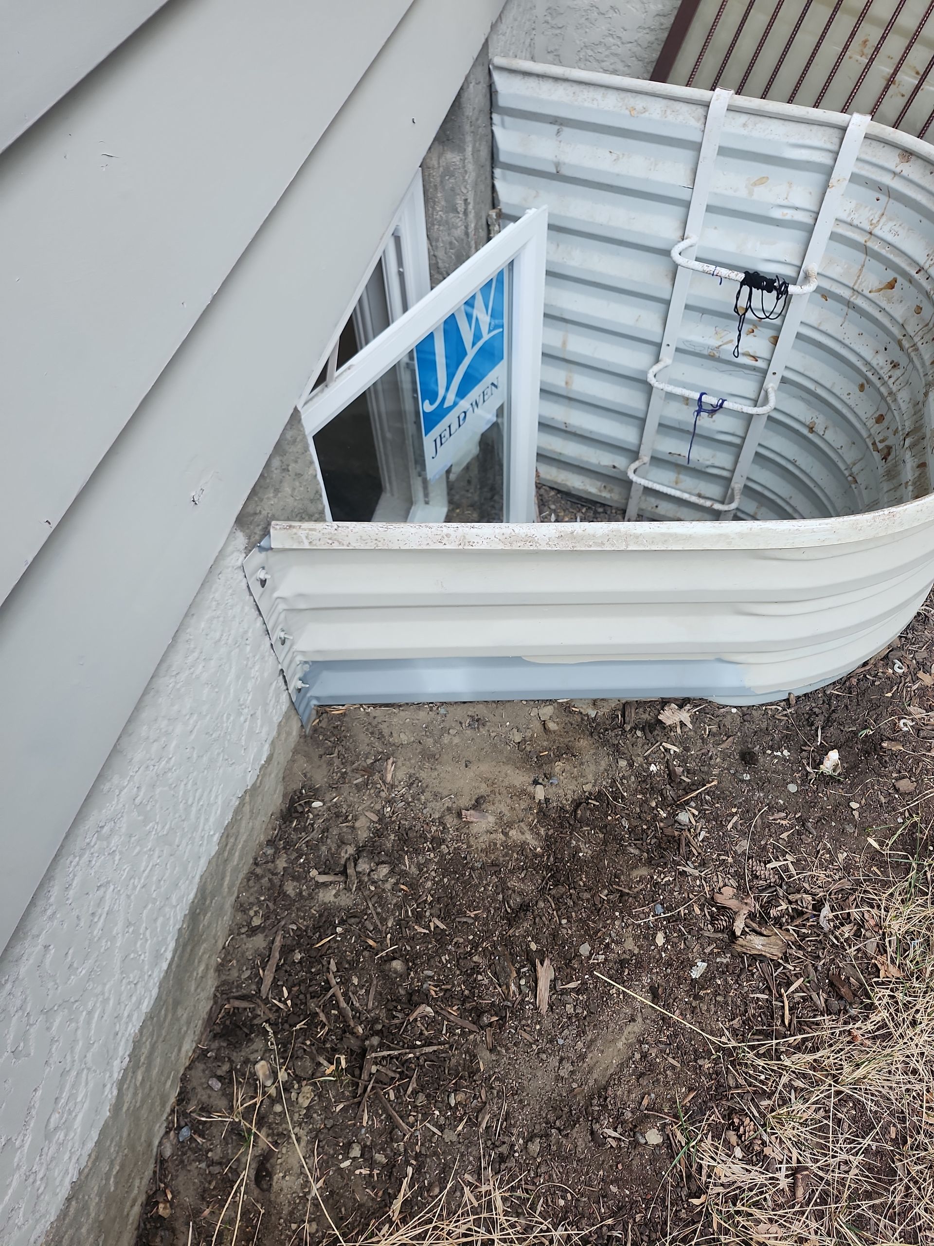 White basement window with a curved well and ladder, next to a building wall.