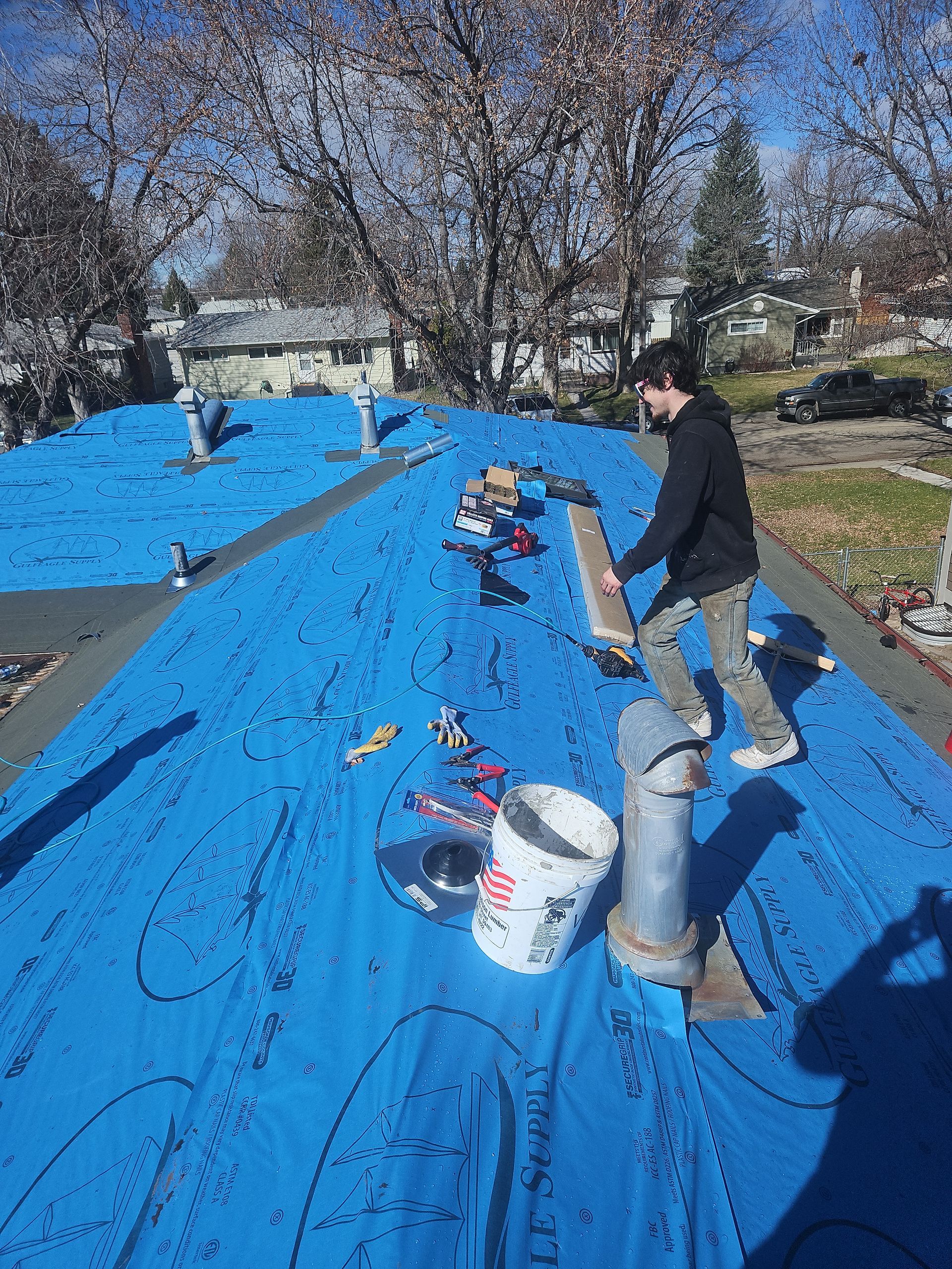 Person on a roof covered in blue tarp, carrying a board near tools and a pipe.