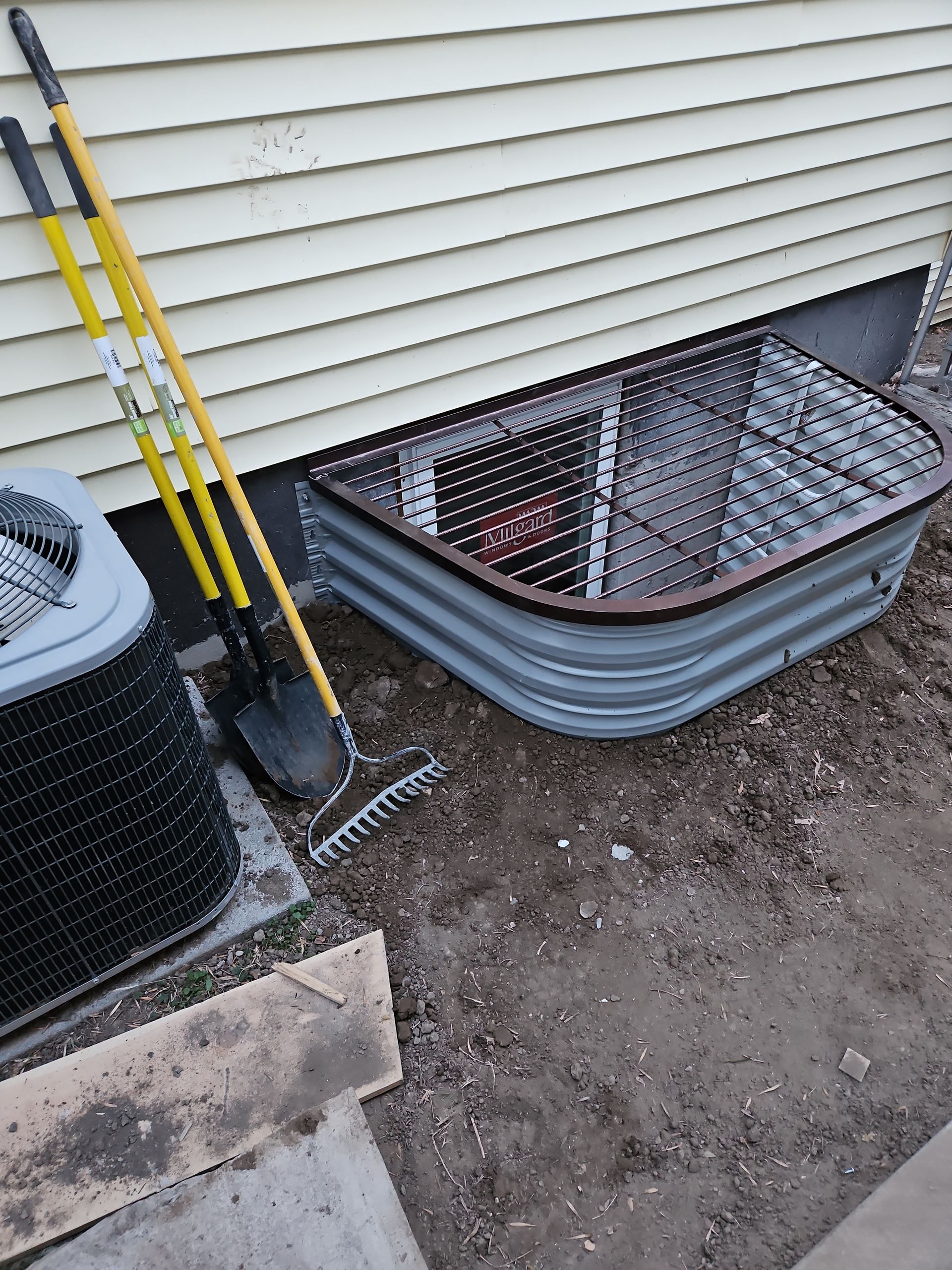 Exterior view: metal window well with mesh cover, next to yellow-handled yard tools and air conditioning unit.