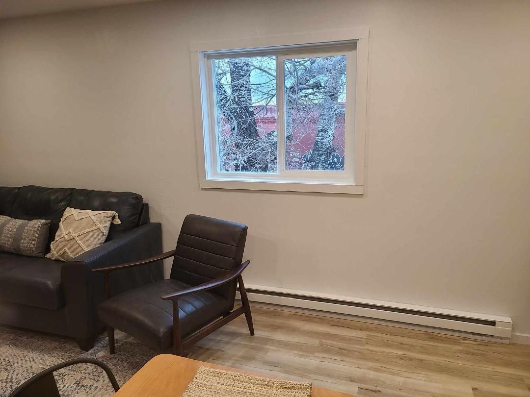 A room with a sofa, chair, and window. Beige walls, wood floor, and a radiator under the window.