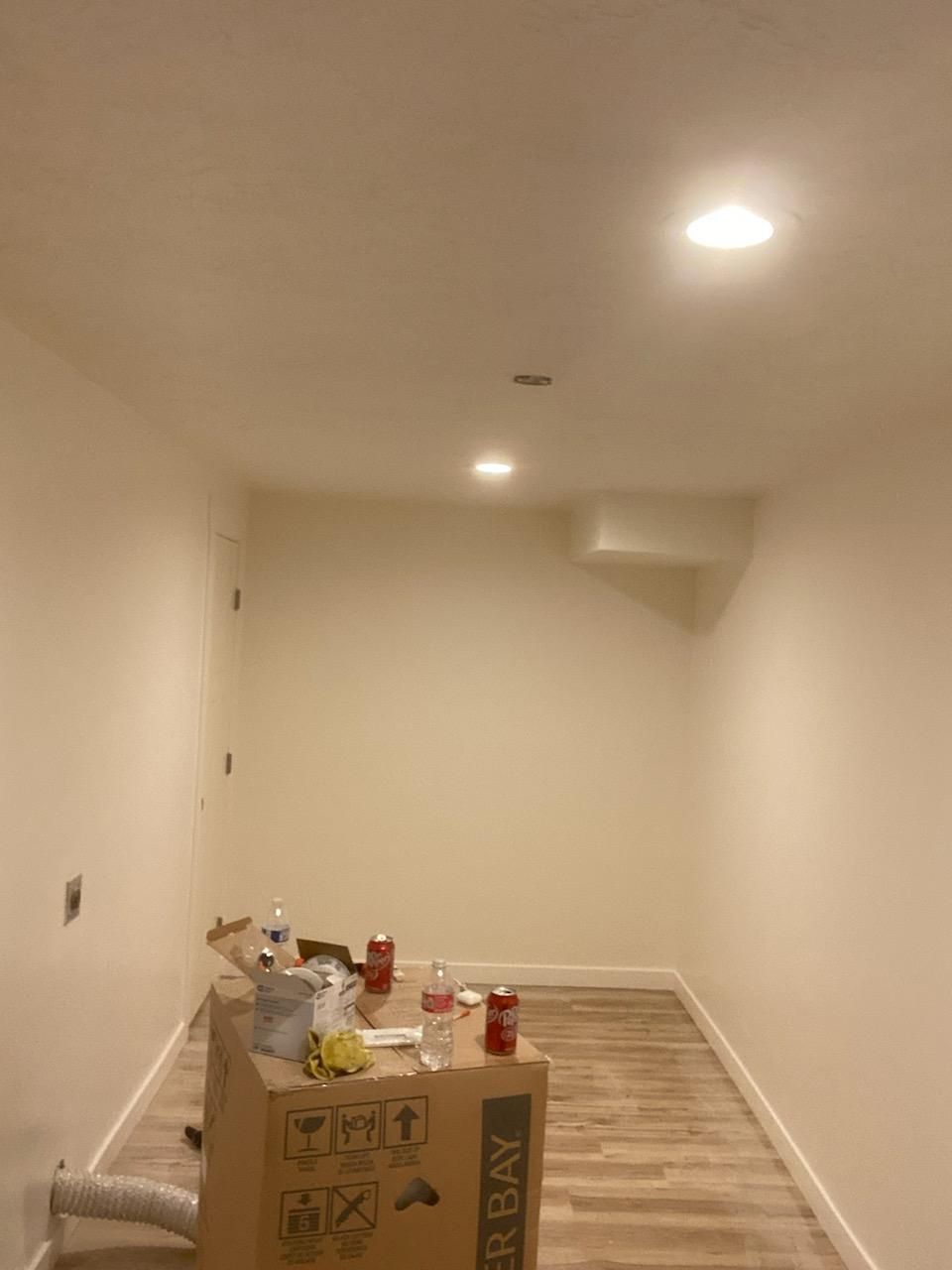Interior of a small, unfinished room with recessed lighting, painted beige walls, and wood-look flooring; a cardboard box sits in the center.