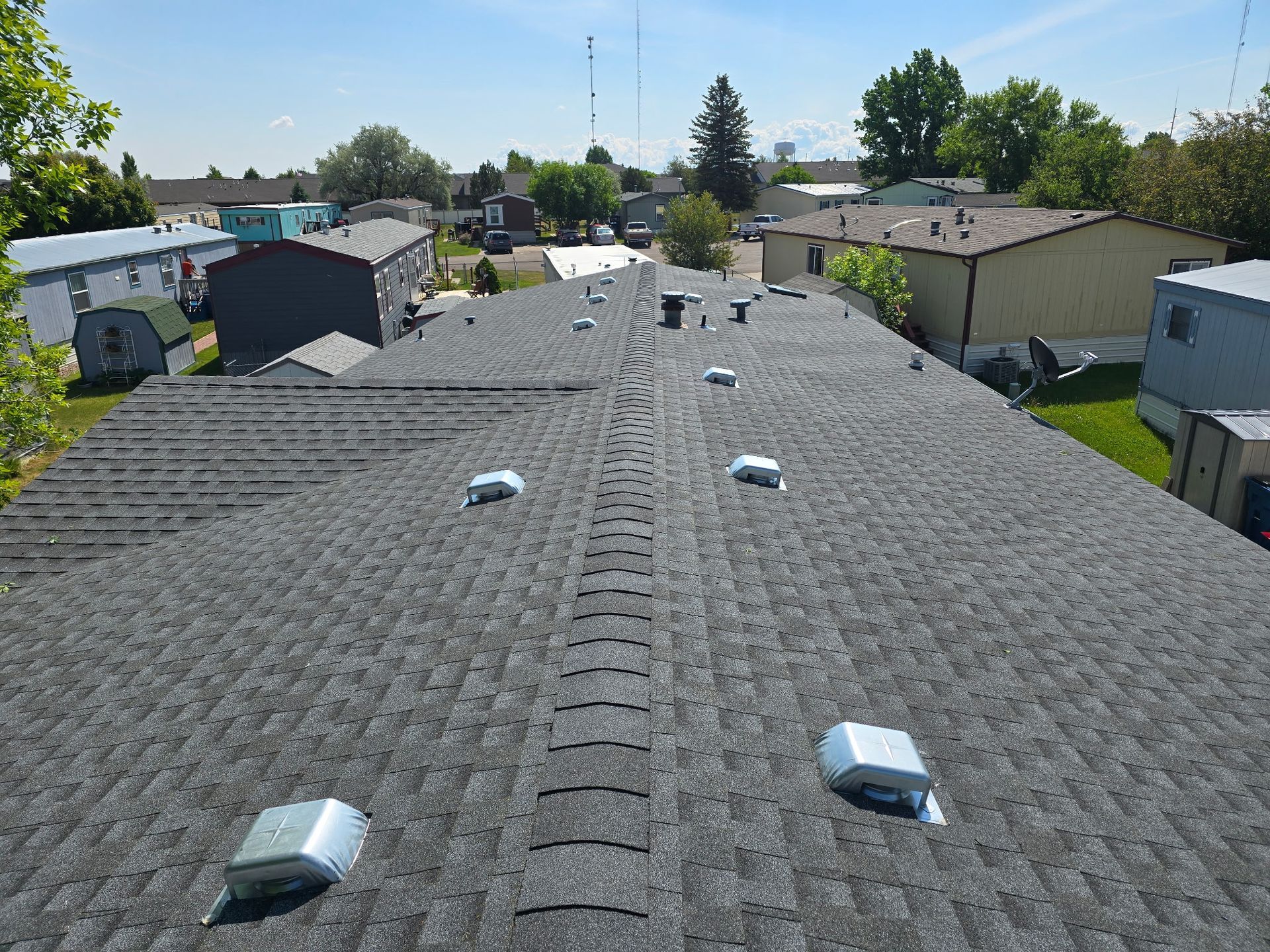 Dark gray shingle roof of a house, vent pipes visible. Suburban setting, blue sky.