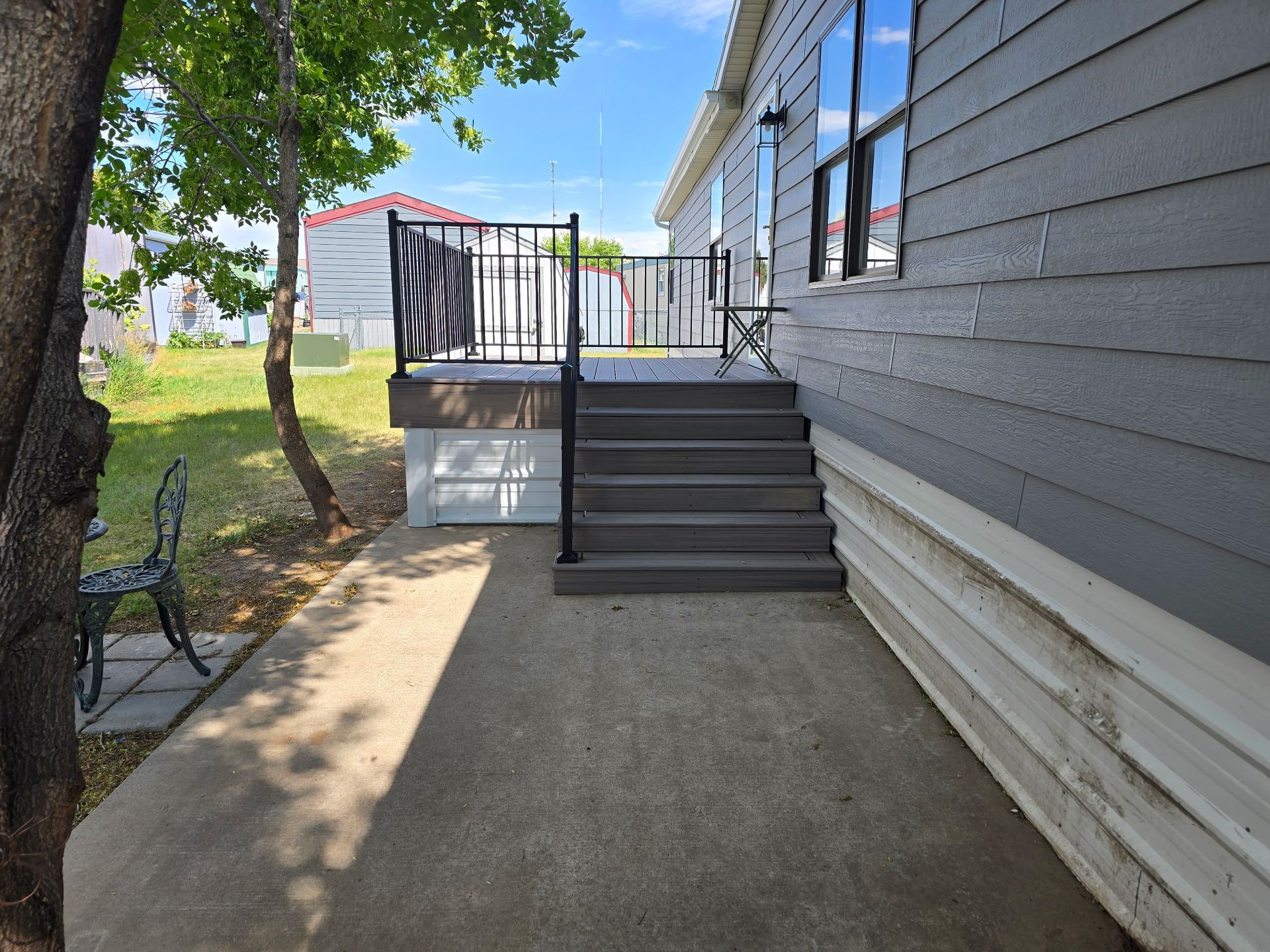 Exterior of a house with steps leading to a deck, black railing, concrete patio, gray siding, and a tree.