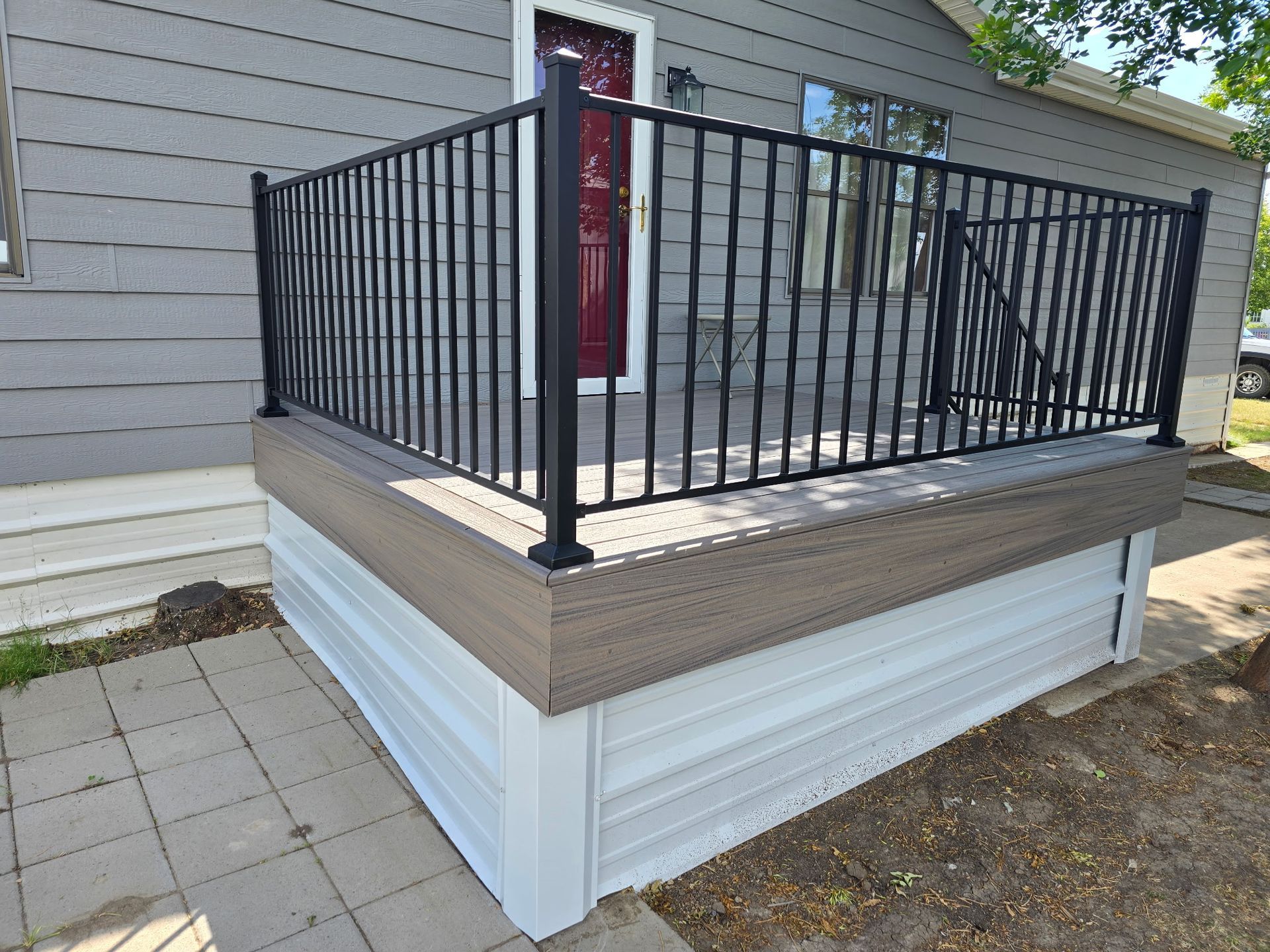 Elevated deck with black railing and light gray siding, on a paved walkway.