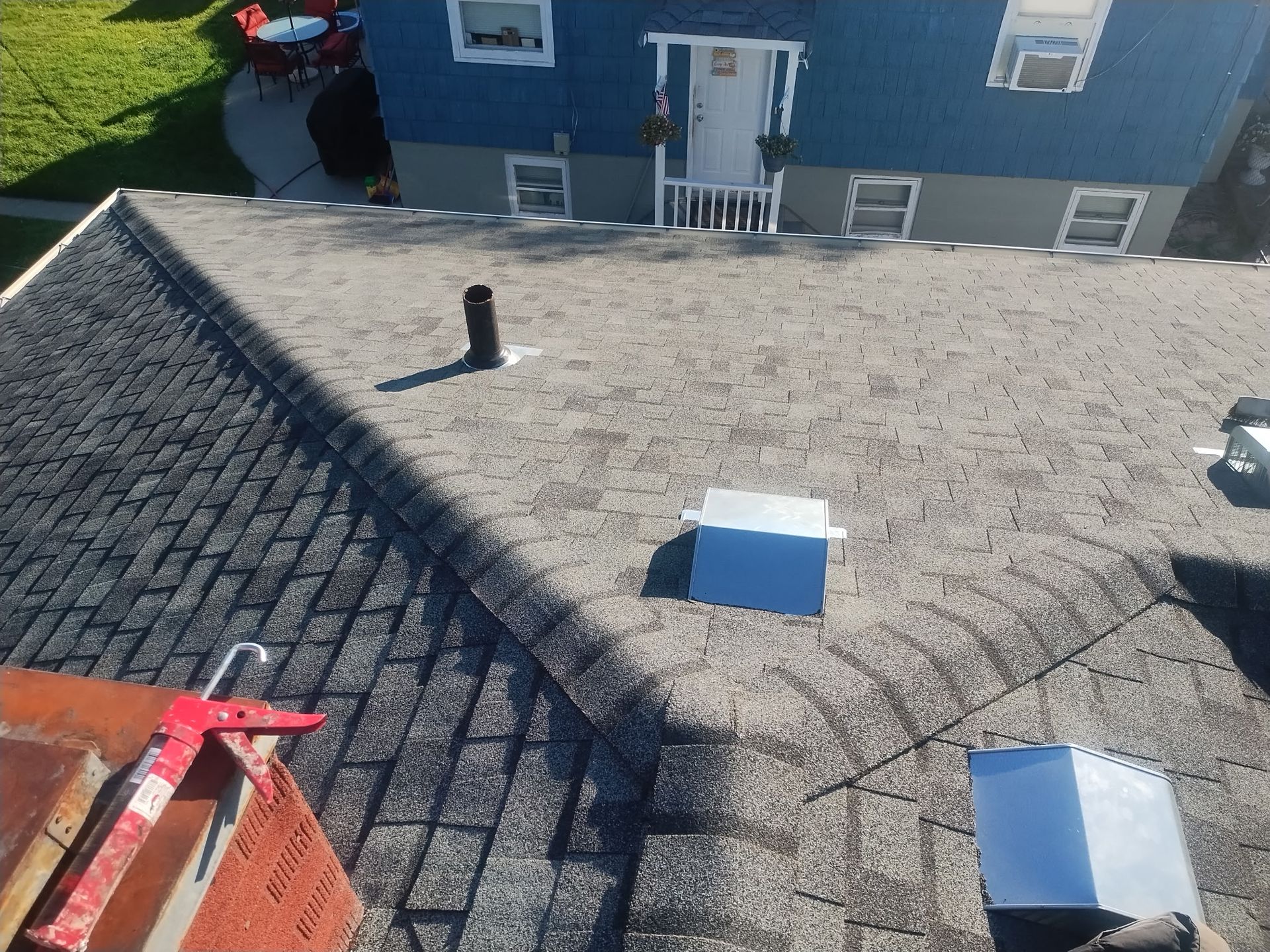 Rooftop with shingles and vents.  House in the background with blue siding.  Bright sunlight.