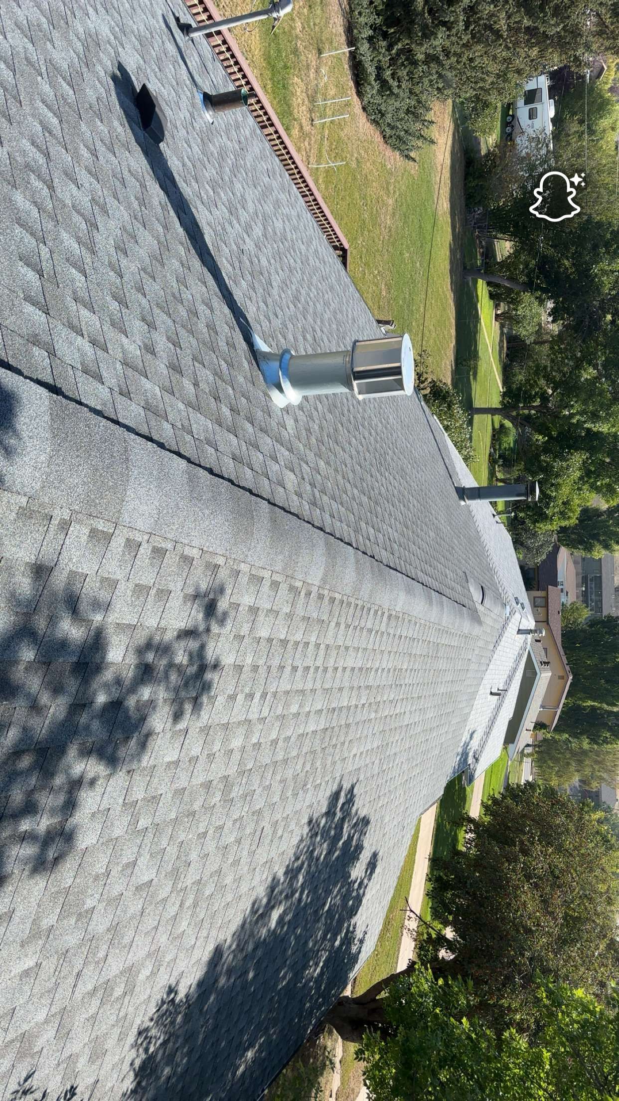 Overhead view of a gray shingled roof with chimneys and trees visible around a sunny day.