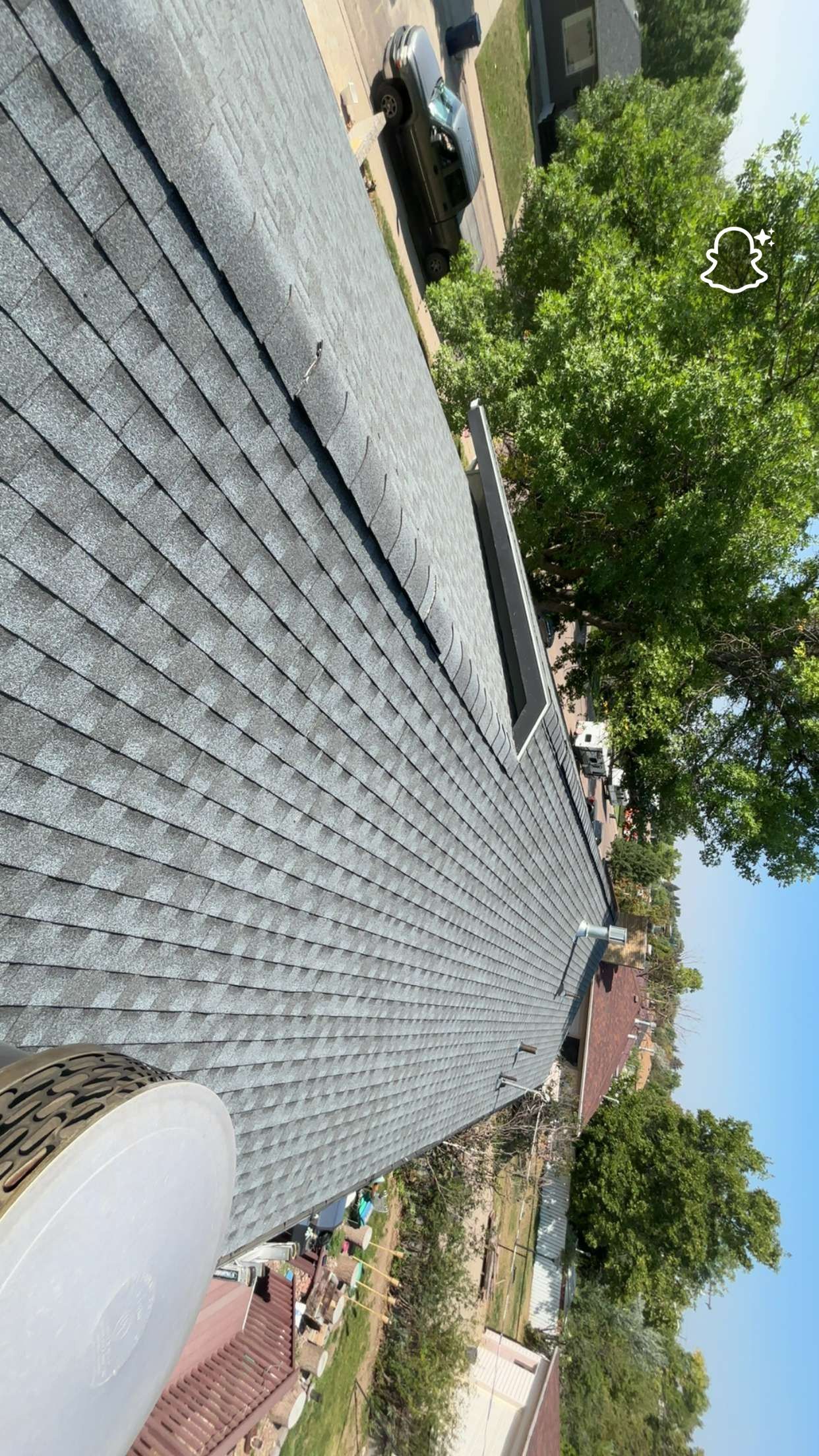 Gray shingled roof of a house, seen from above. Trees and a vehicle are nearby.