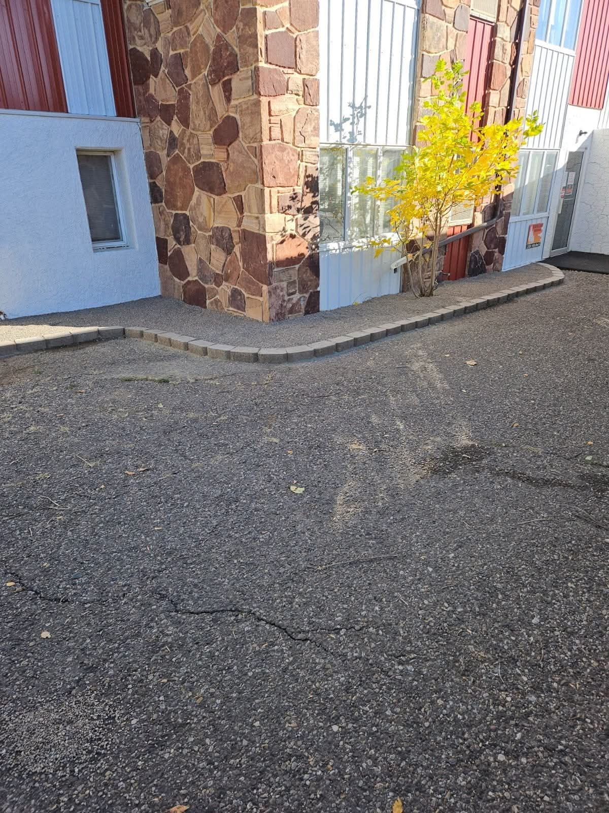 Corner of building with brown and red stone facade, asphalt in foreground, small yellow tree.