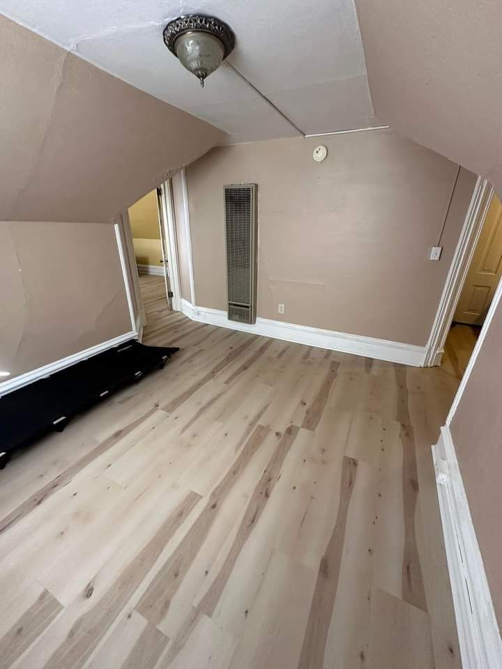 Room with light wooden floors, tan walls, and a ceiling light. A hallway and door are visible.