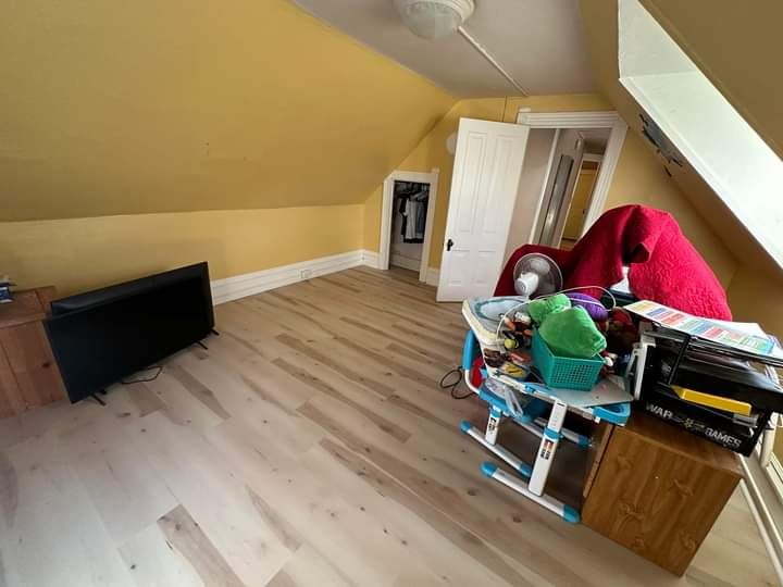 Yellow-walled attic room with angled ceiling, light wood floor, TV, and cluttered table near a window.