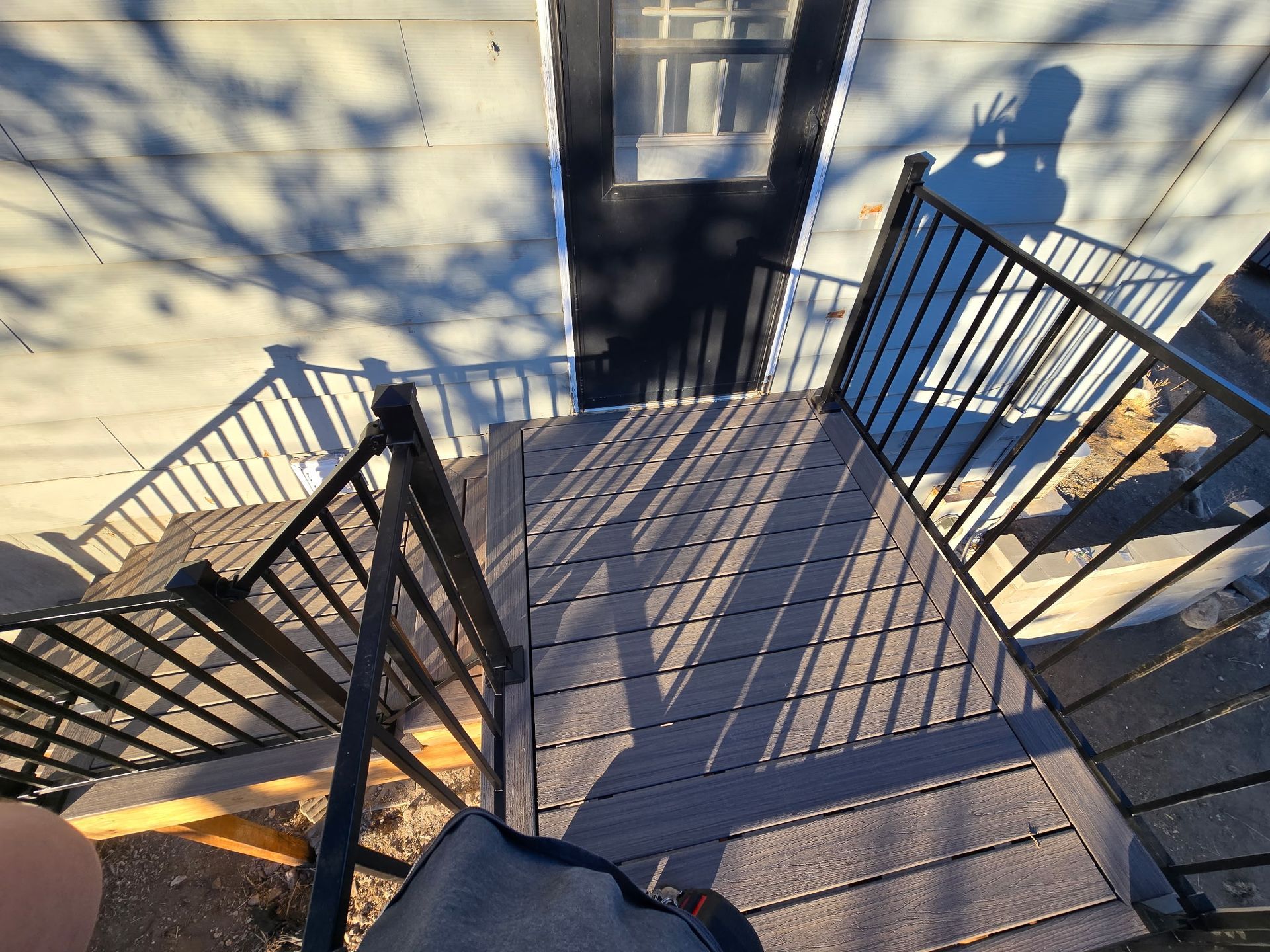 Top-down view of outdoor stairs leading to a door; black railings, wooden steps, and long shadows cast by sunlight.