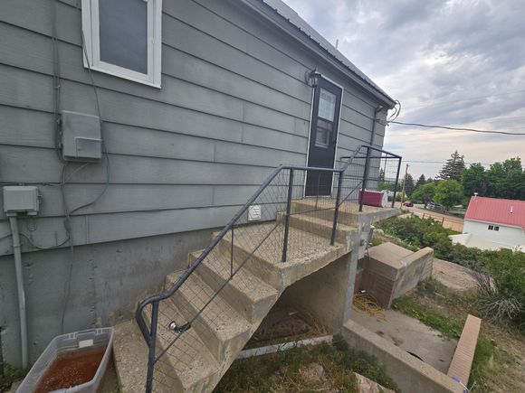 Gray house exterior with concrete steps leading to a door. Handrails and overgrown grass. Cloudy sky.