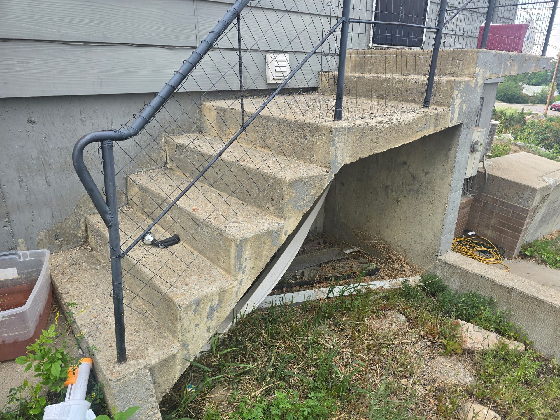 Concrete exterior stairs with a black metal handrail leading to a door. Underneath the stairs is overgrown with weeds.