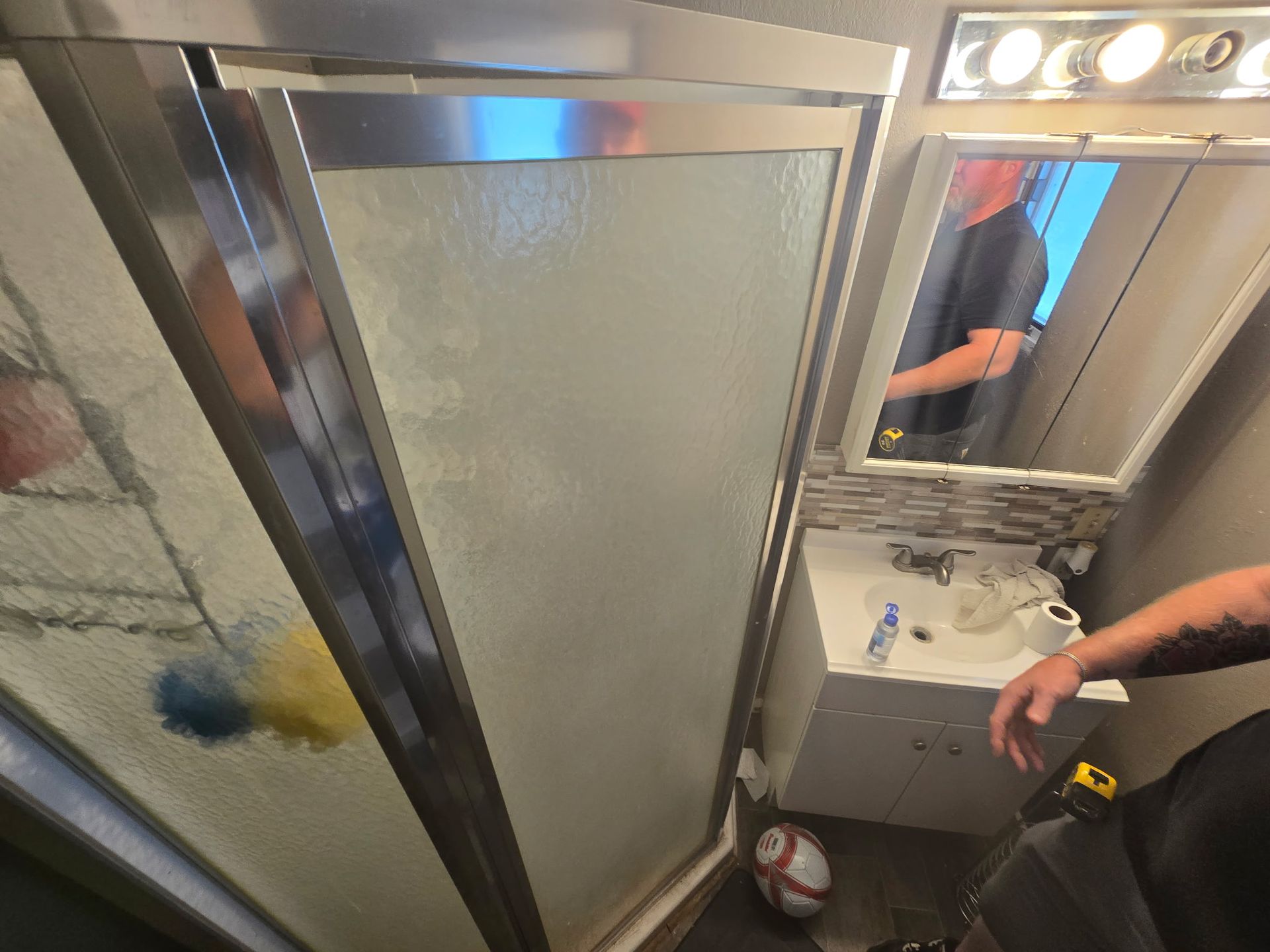 Shower stall with glass door, small vanity with mirror, and a person in the mirror.