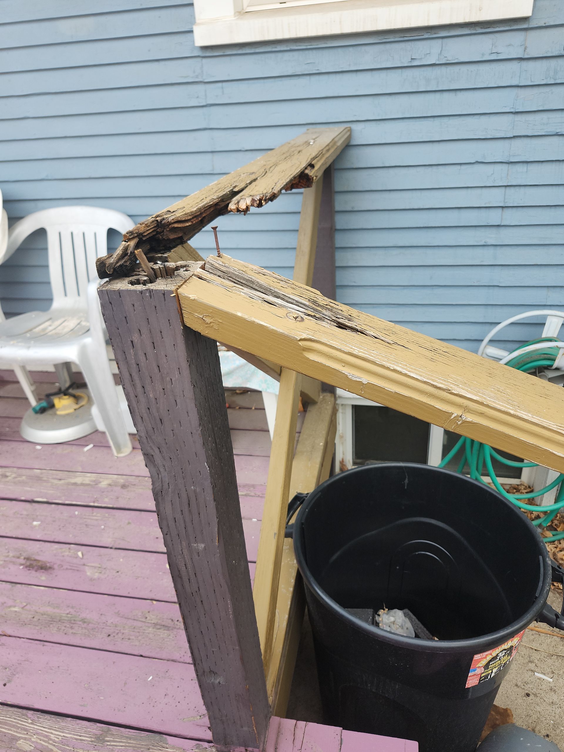 Damaged deck railing, partially broken, against blue siding and purple deck. Black bucket in view.
