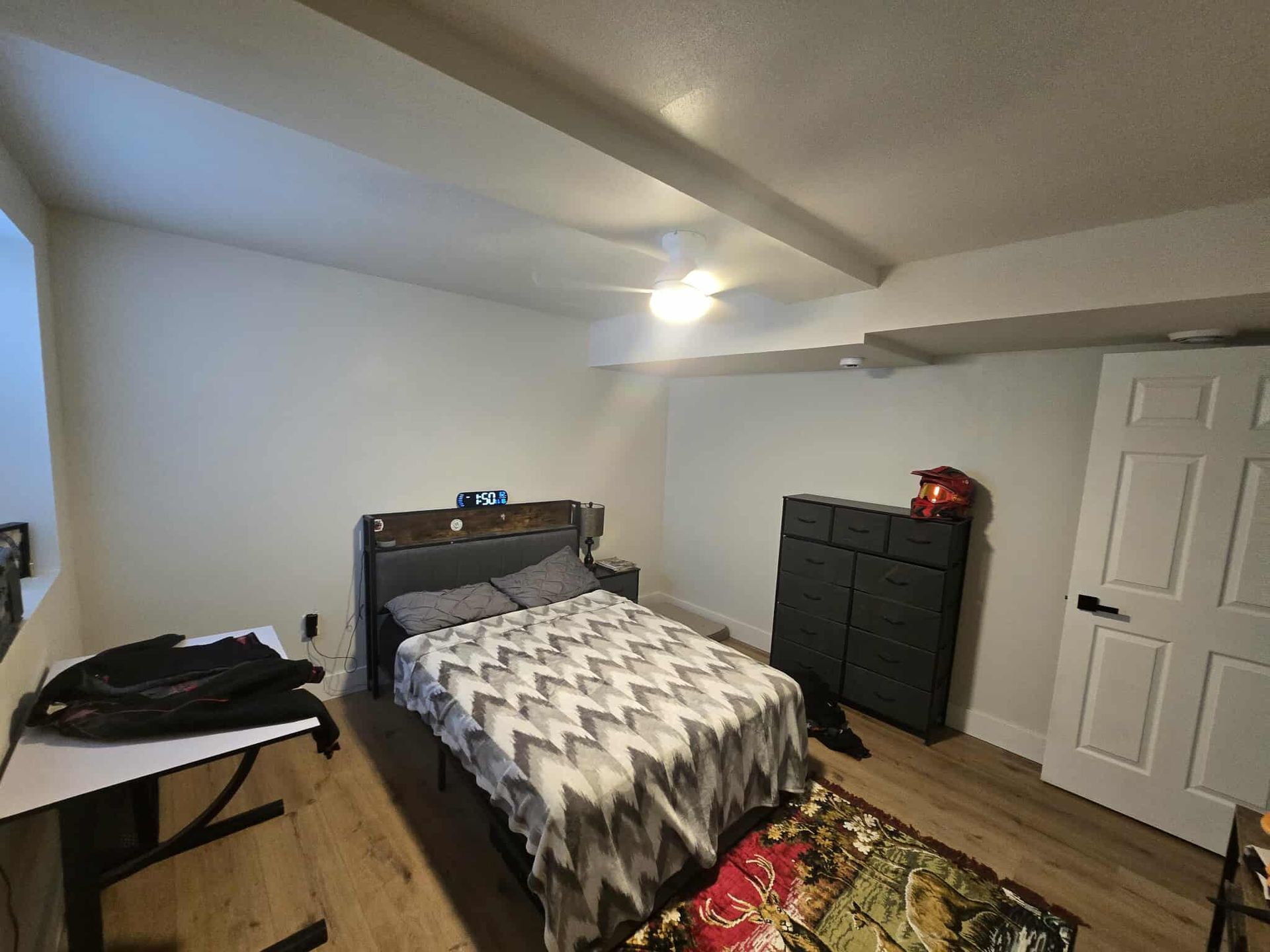 Bedroom with bed, desk, dresser, and door.  White walls, wood floor, and a small area rug.