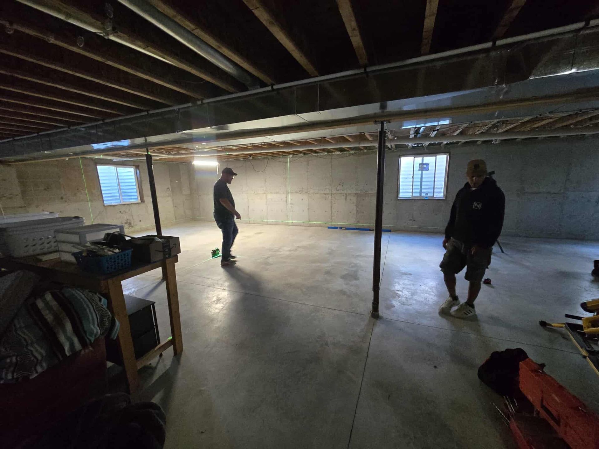 Two men standing in an unfinished basement with support beams and concrete walls.