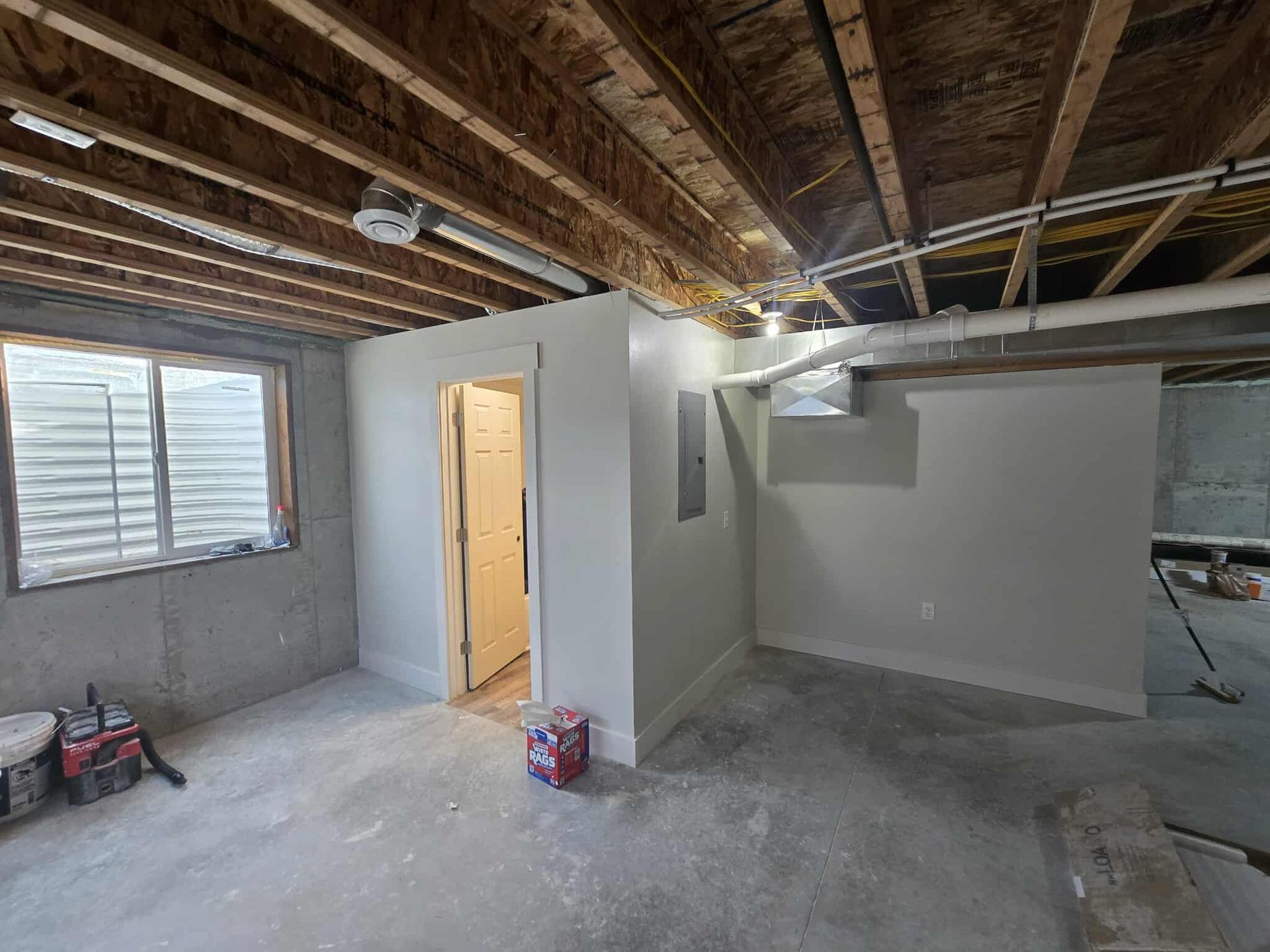 Basement interior with exposed ceiling beams, unfinished walls, a door, and a window.