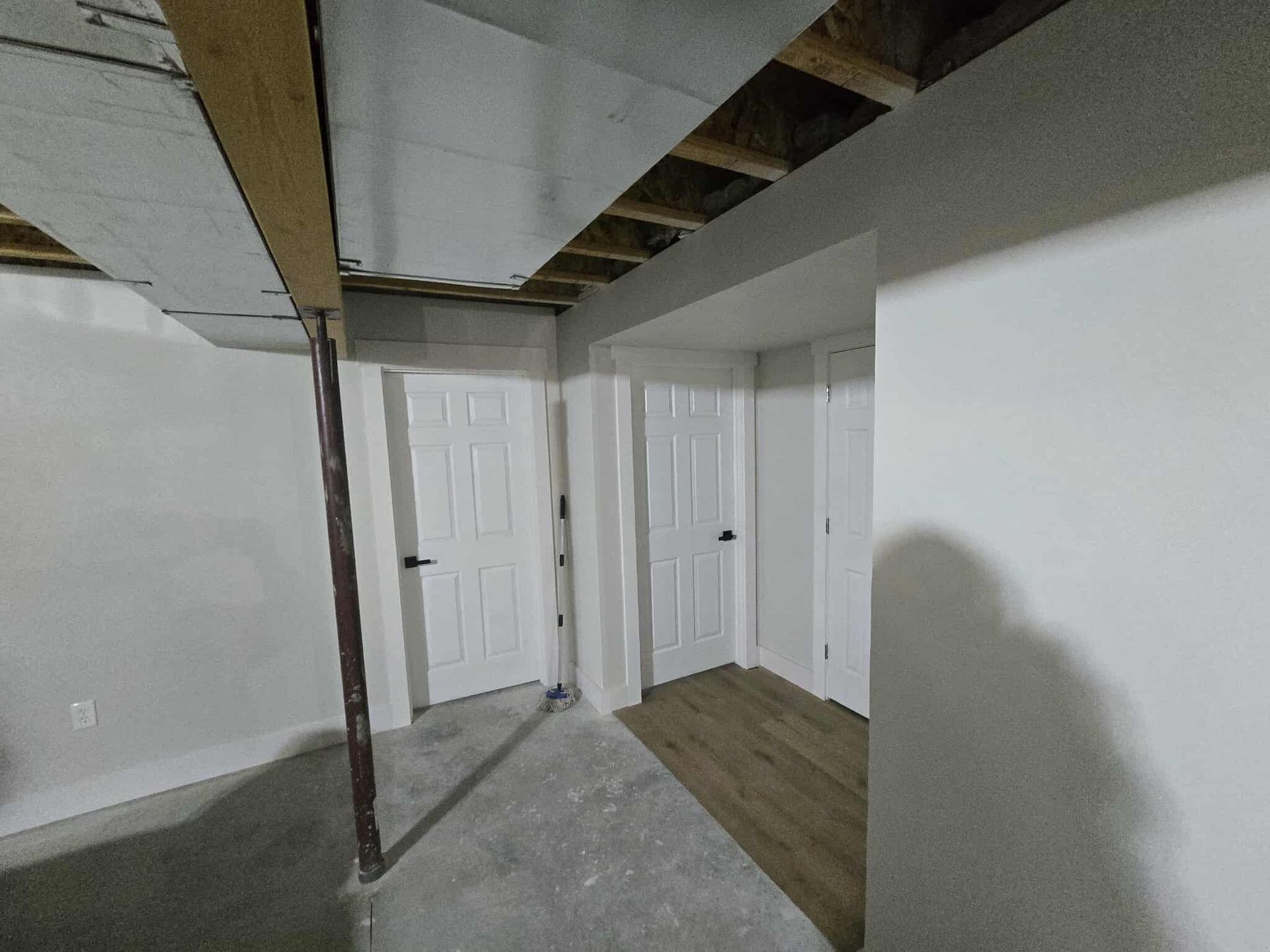 Basement hallway with two white doors, support beam, exposed ceiling beams, and concrete floor.