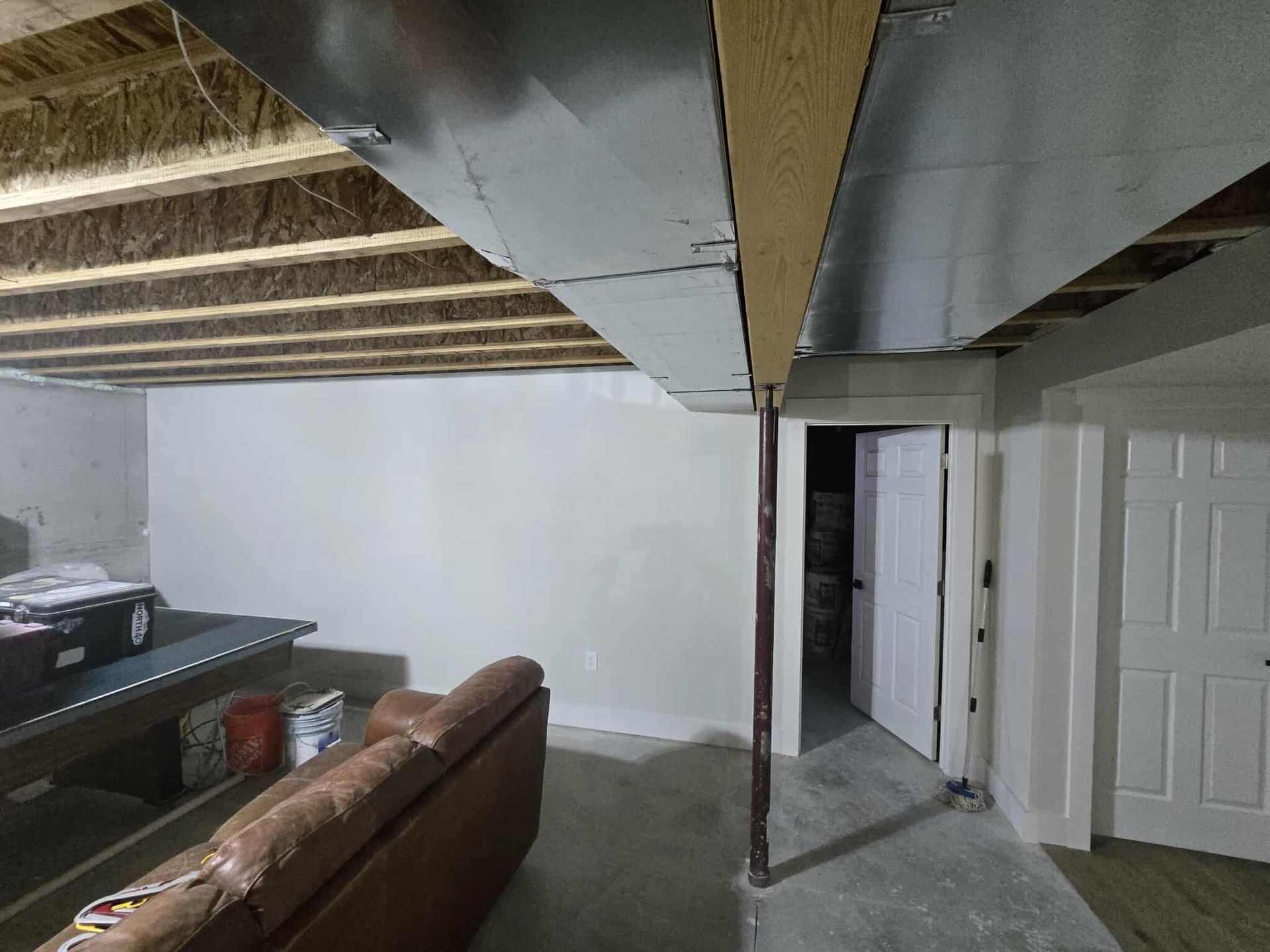 Basement interior with exposed ceiling beams, ductwork, and partially finished walls. A couch and open doorway are visible.