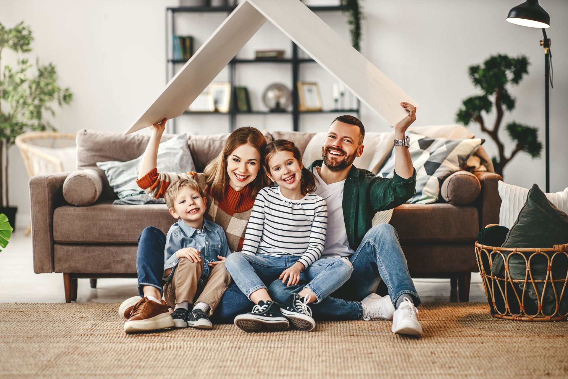 Une famille de quatre personnes, souriante, assise sur un tapis dans un salon, soutenant un toit en carton.
