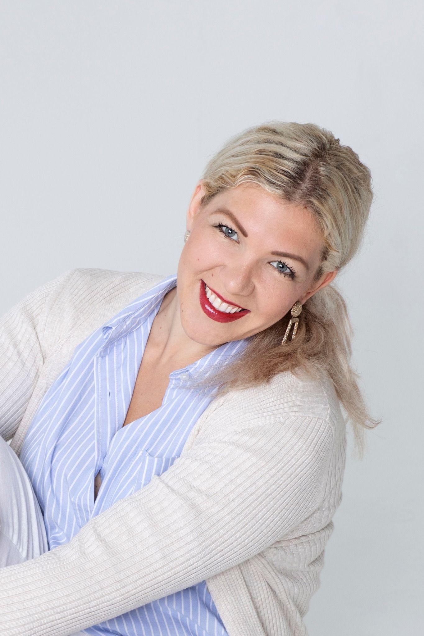 Femme blonde, souriant largement, portant une chemise à rayures bleues et un cardigan crème.