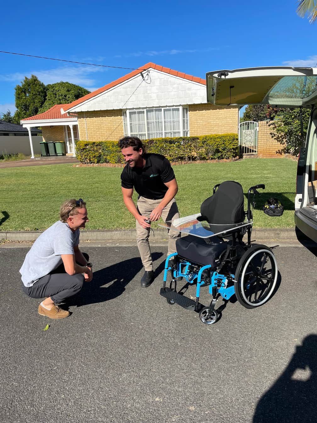 Two men are standing next to a wheelchair on the side of the road.