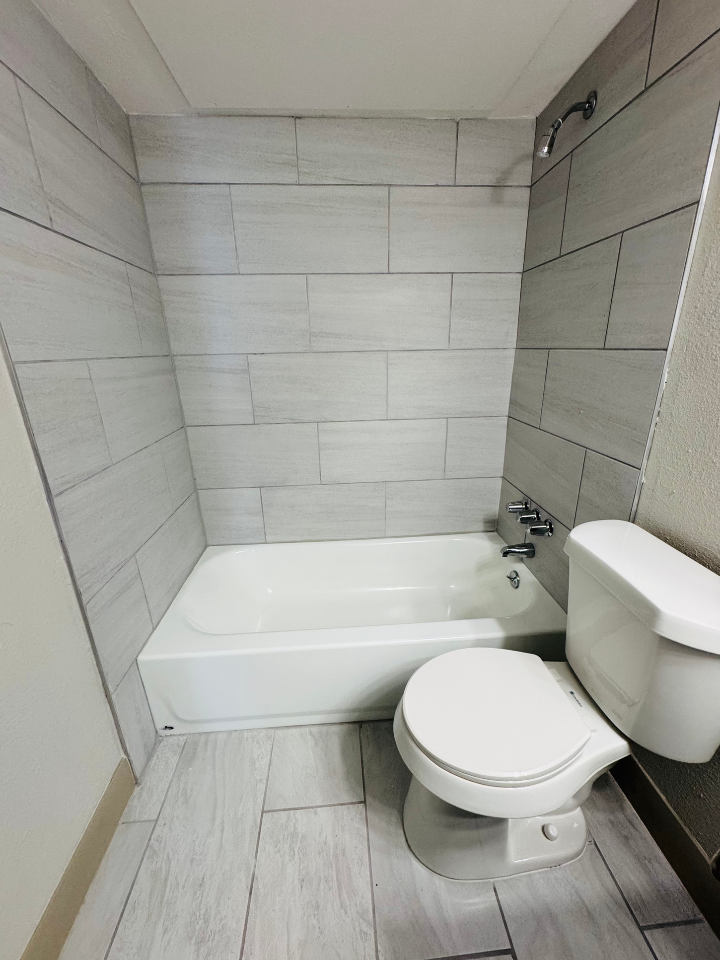 A white bathroom with a bathtub, toilet, and light gray tile walls and floor.