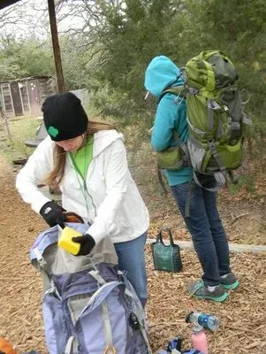 Two hikers packing gear on a wooded trail, one wearing a large green backpack.