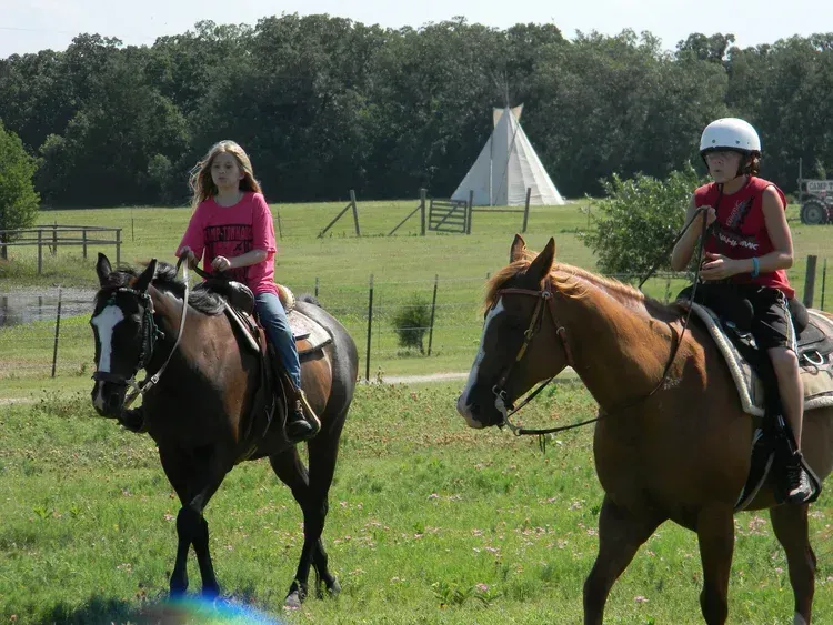 Two horseback riders in a grassy field, with a teepee and trees in the background.