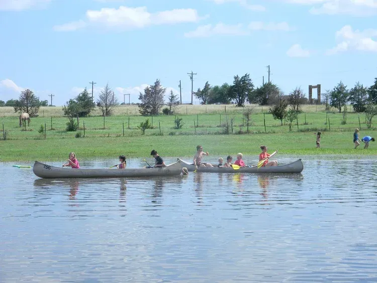People paddling a long canoe across a calm lake in a grassy park under a blue sky