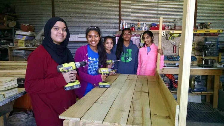 Four people stand behind a wooden table in a workshop, holding stacked items and smiling.