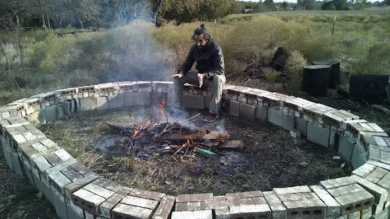 Person sitting by a small fire in a circular stone fire pit outdoors.