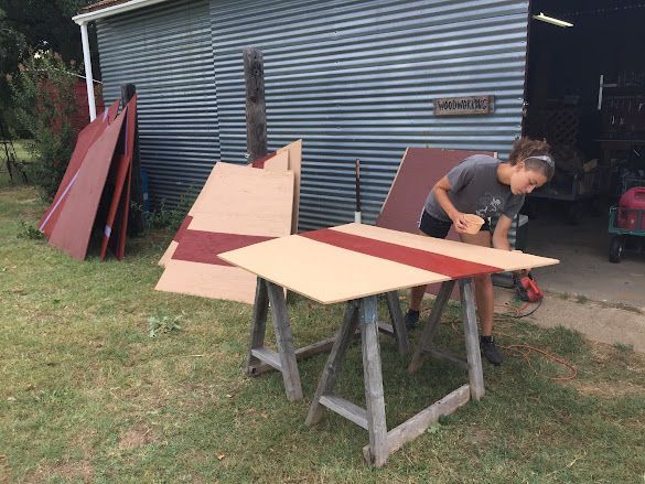 Person working on a red-and-beige wooden panel on sawhorses outside a shed