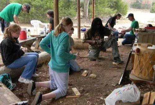 People kneel and work together outdoors around drums in a shaded wooded area.