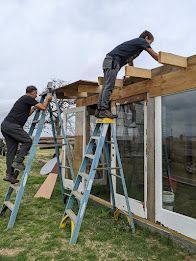 Two people repairing a wooden roof frame on ladders beside a house