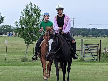 Two people wearing helmets ride horses side by side on a grassy field.