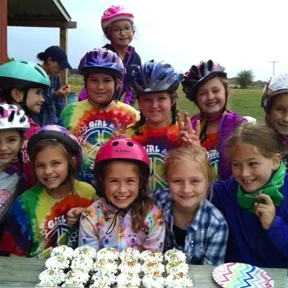 Children in helmets smiling around a cake outdoors, with a girl in a pink helmet at the center