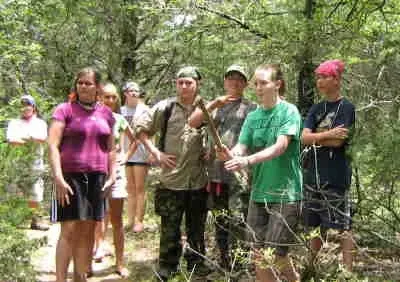 Group of hikers standing on a wooded trail, some holding walking sticks, with a bright pink shirt and green shirt visible