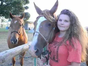 Girl standing beside two brown horses by a wooden fence outdoors