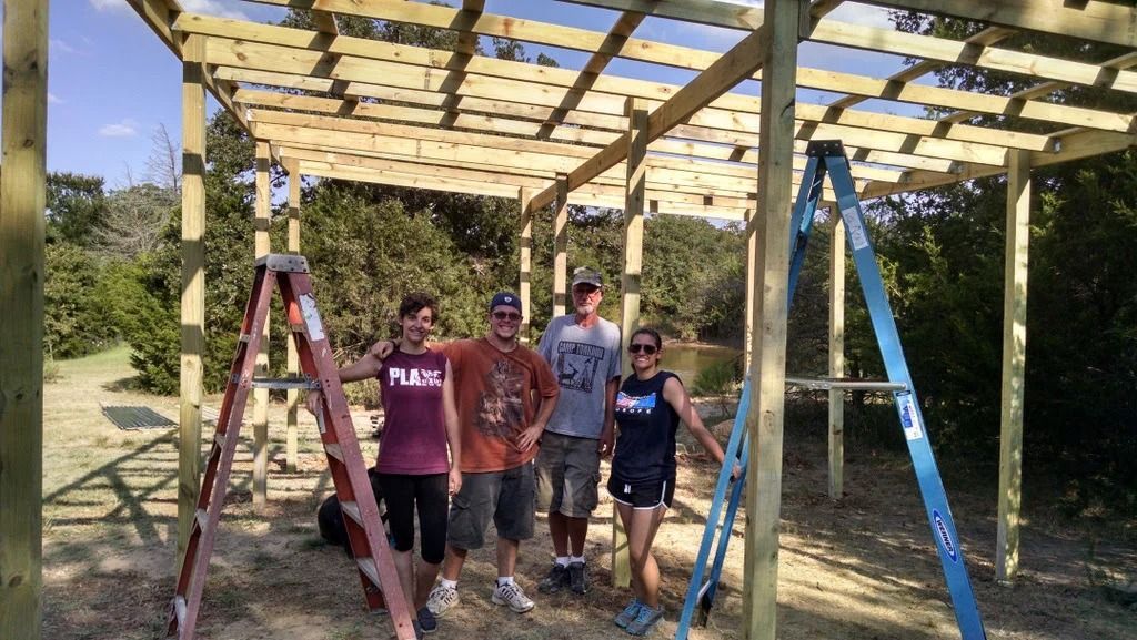 Group standing under a partially built wooden pergola on a sunny outdoor site.