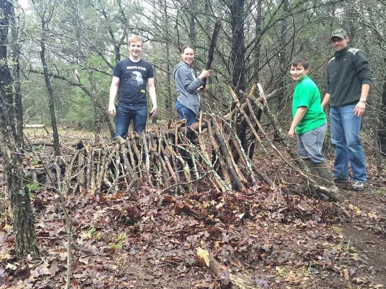 Four people standing beside a handmade stick fence in a wooded area on a muddy trail