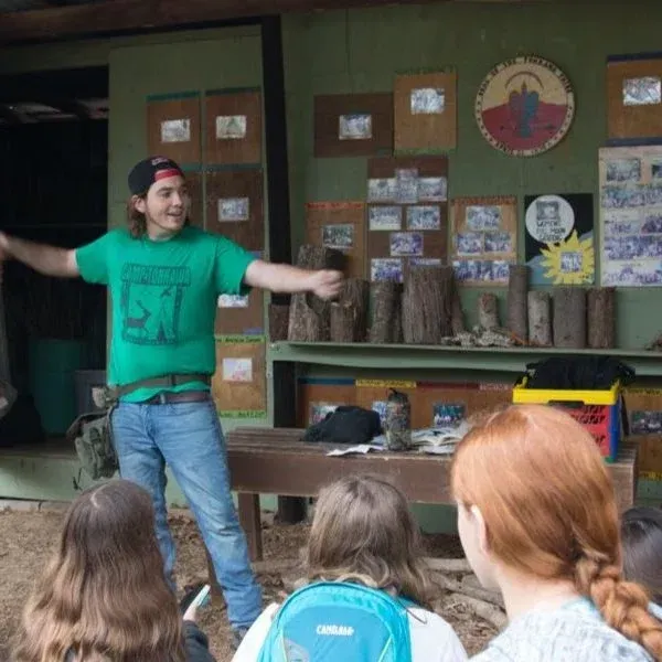 Person in a green shirt gesturing in front of a group seated outdoors near a cabin.
