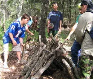 People standing around a campfire pile in a wooded area, examining stacked logs.