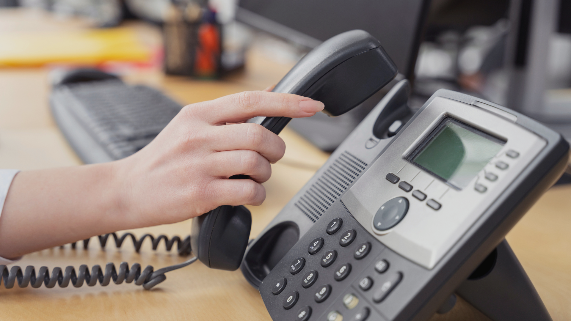 Hand picking up a black landline phone. The phone rests on a desk with a keyboard visible.